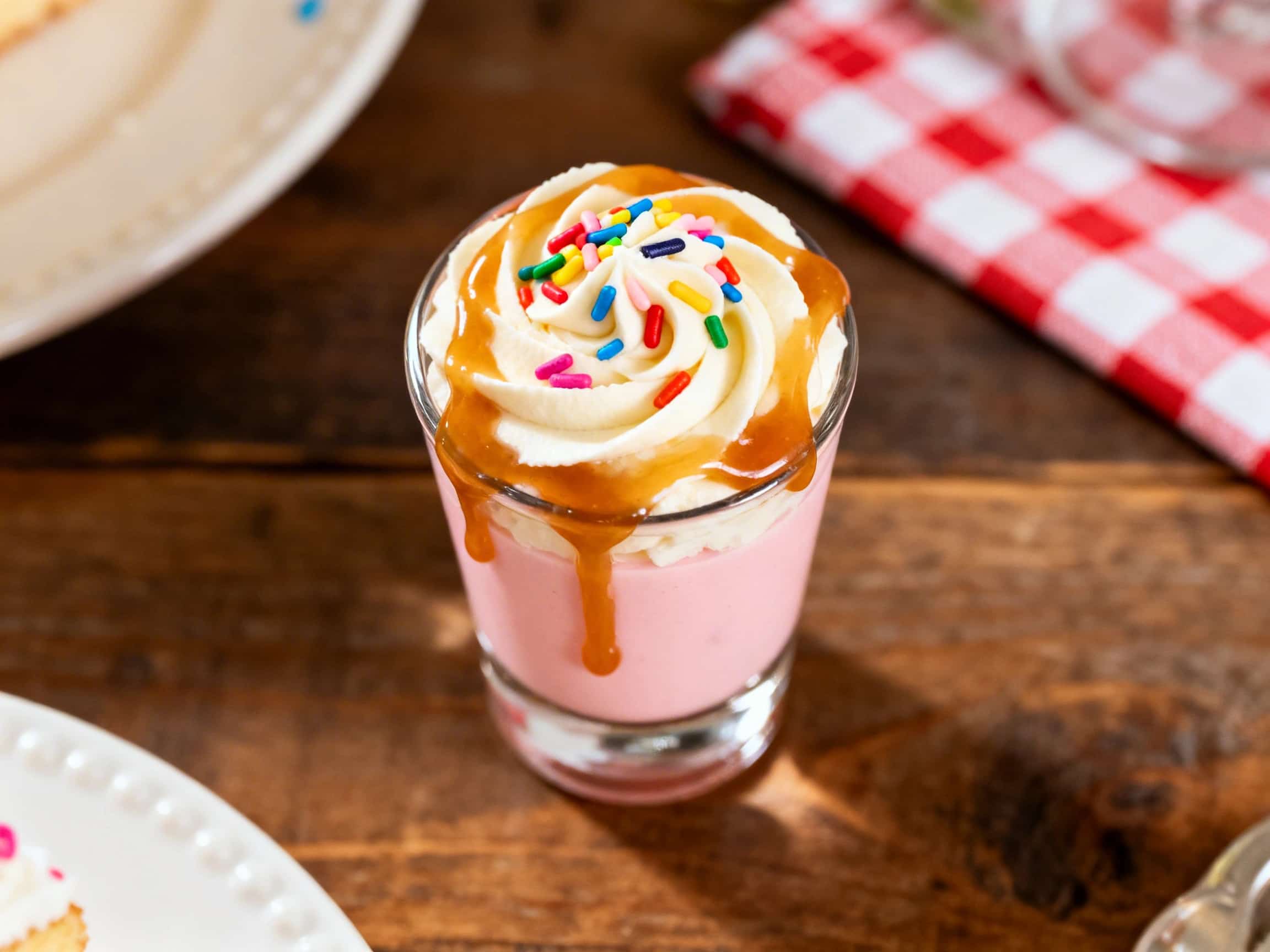 A single Birthday Cake Vodka Shot in a clear, small shot glass, presented against a soft, festive background. The creamy, pale yellow liquid fills the glass, and the rim is generously coated with bright, multicolored sprinkles. The overall presentation is inviting and celebratory, capturing the essence of a fun, dessert-themed beverage.
