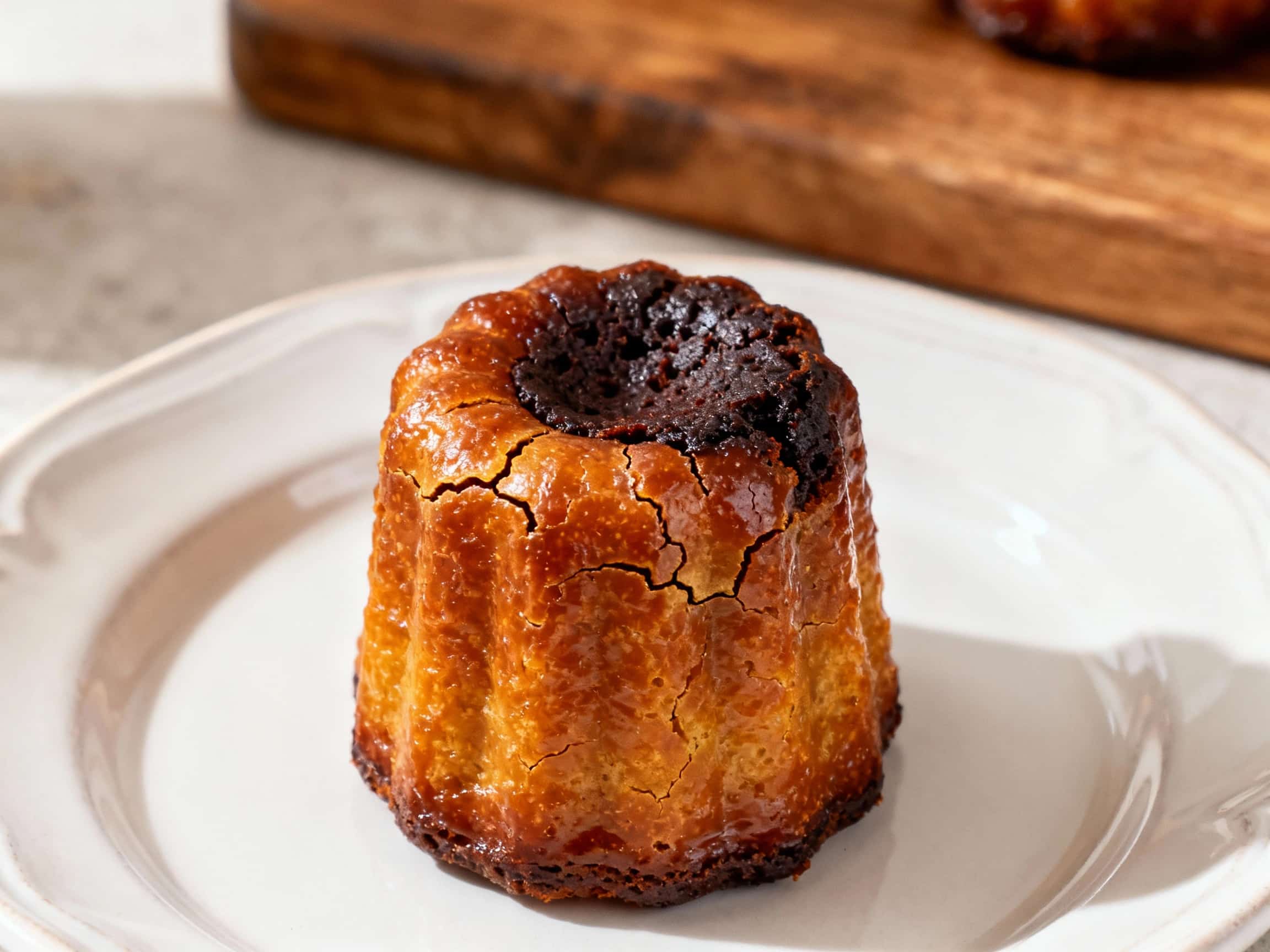 Golden-brown Bordeaux cannelés with a tall, caramelized crust and a custardy, dark interior sit attractively arranged on a rustic wooden board, ready to be enjoyed.