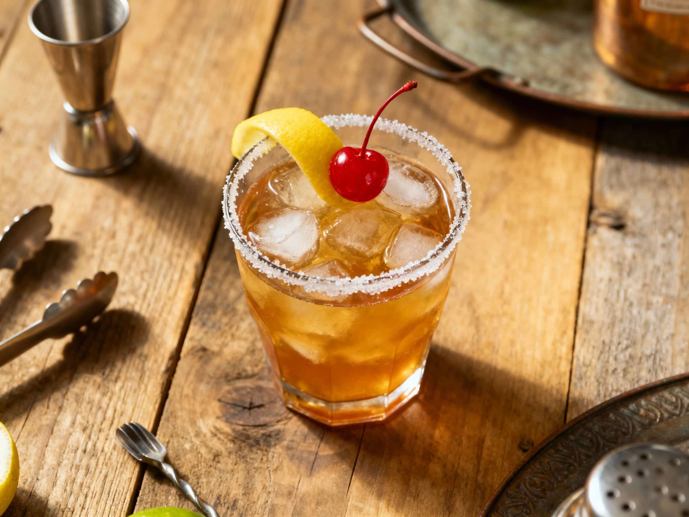 A frothy, golden-hued Classic Whiskey Sour cocktail in a crystal coupe glass, elegantly garnished with a lemon wheel and a bright red maraschino cherry against a soft, dark background. The drink features a thick, creamy white foam top, with a few dashes of aromatic bitters creating a beautiful contrast. The inviting atmosphere suggests a sophisticated, classic cocktail experience.