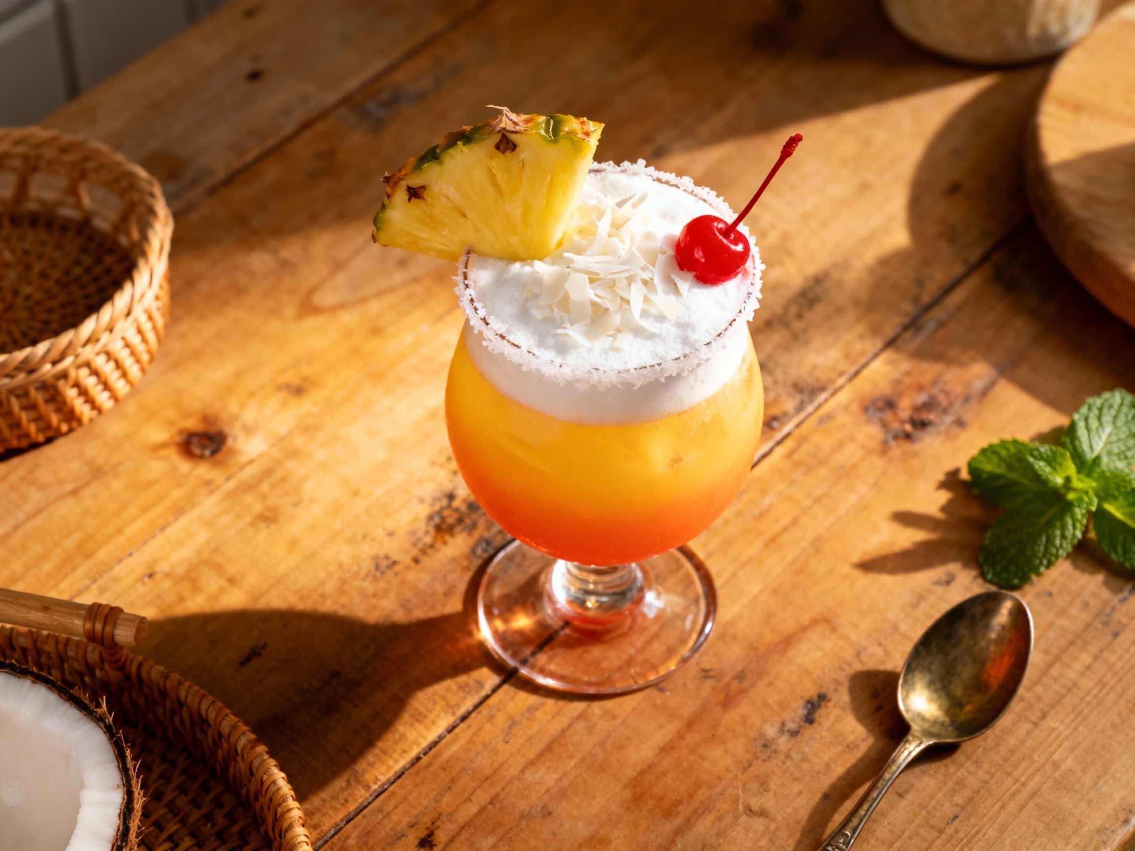 A vibrant orange and yellow layered cocktail with a slice of pineapple and a cherry garnish sits tall in a glass, evoking a tropical sunrise.