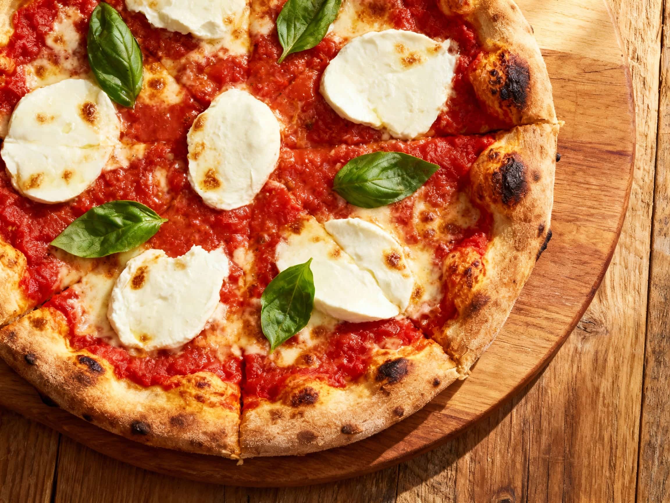 A stunning, freshly baked New York-style Margherita pizza sits on a wooden pizza peel. The pizza boasts a classic thin crust with a beautifully golden and slightly charred edge, indicative of high-heat baking. The surface is adorned with vibrant red tomato sauce, creamy melted fresh mozzarella, and aromatic bright green basil leaves. The cheese has achieved a delightful bubbly texture with pockets