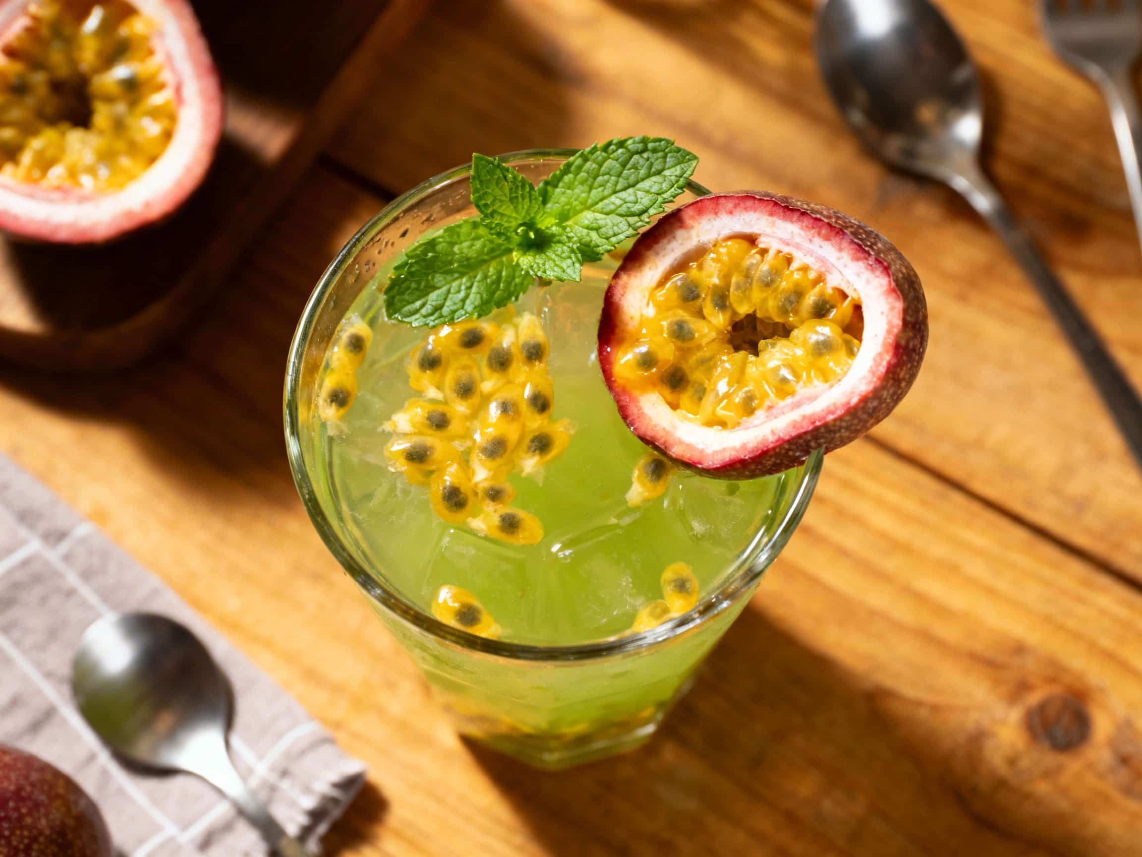 A crystal-clear glass of brightly colored iced green tea, brimming with visible passionfruit pulp and distinctive black seeds, sits elegantly on a wooden surface outdoors. A fresh sprig of mint garnishes the rim, with more mint leaves artfully integrated into the drink. The glass shows condensation, suggesting its chilled refreshment, against a softly blurred backdrop of tropical greenery and a tr