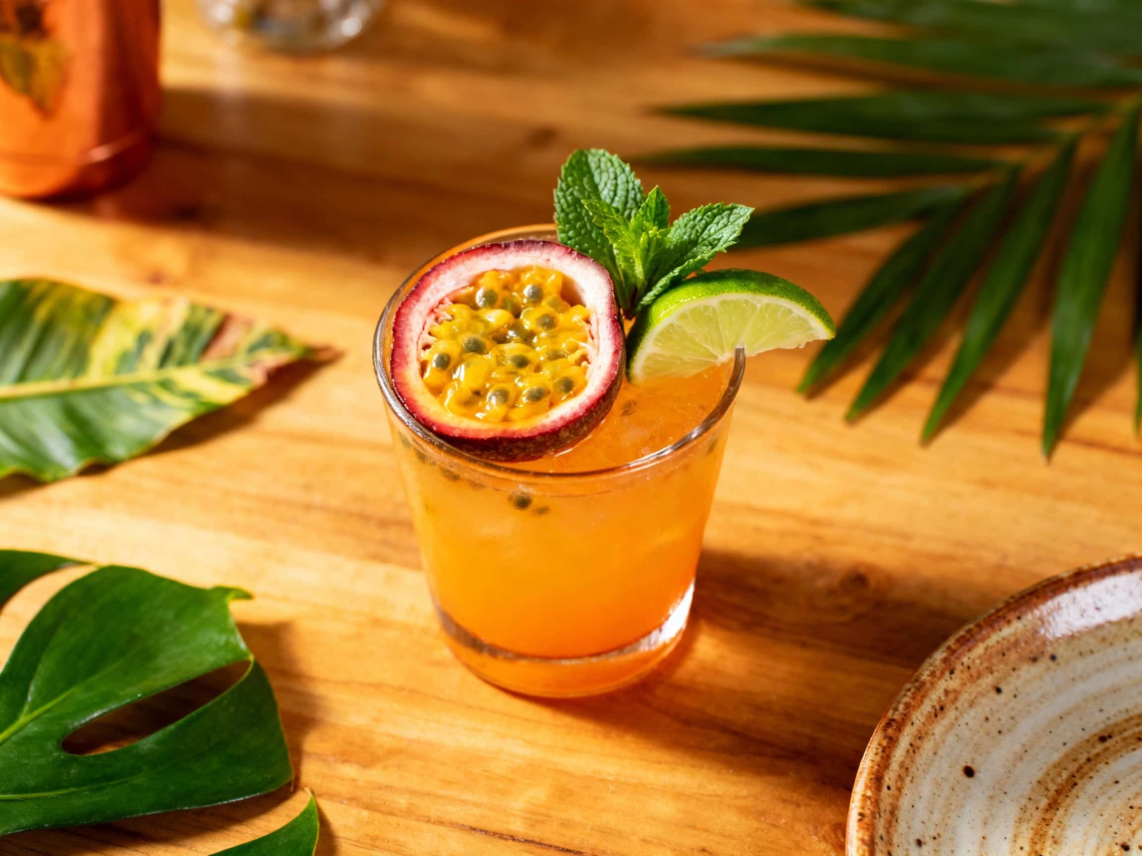 A refreshing Passionfruit Whiskey Smash cocktail in a rocks glass, showcasing a vibrant golden color with ice. It's garnished with a fresh green mint sprig and a slice of passionfruit, suggesting a tropical and inviting beverage perfect for a warm day. The drink appears cool and invigorating, presented against a softly blurred, bright background.