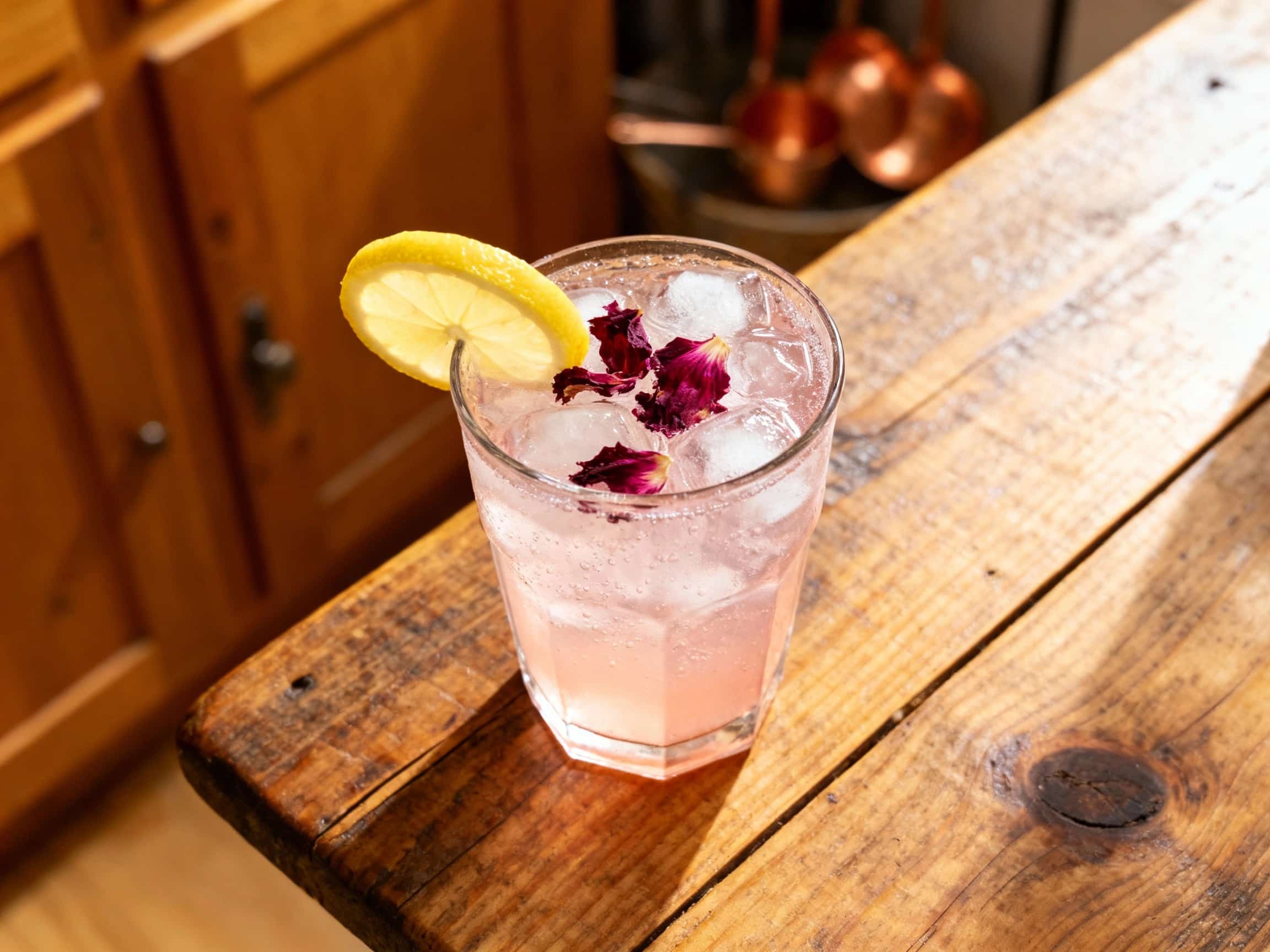 A tall, chilled glass of pink hibiscus gin fizz sits on a white marble surface, garnished with a fresh flower and a sprig of mint, catching the sunlight.