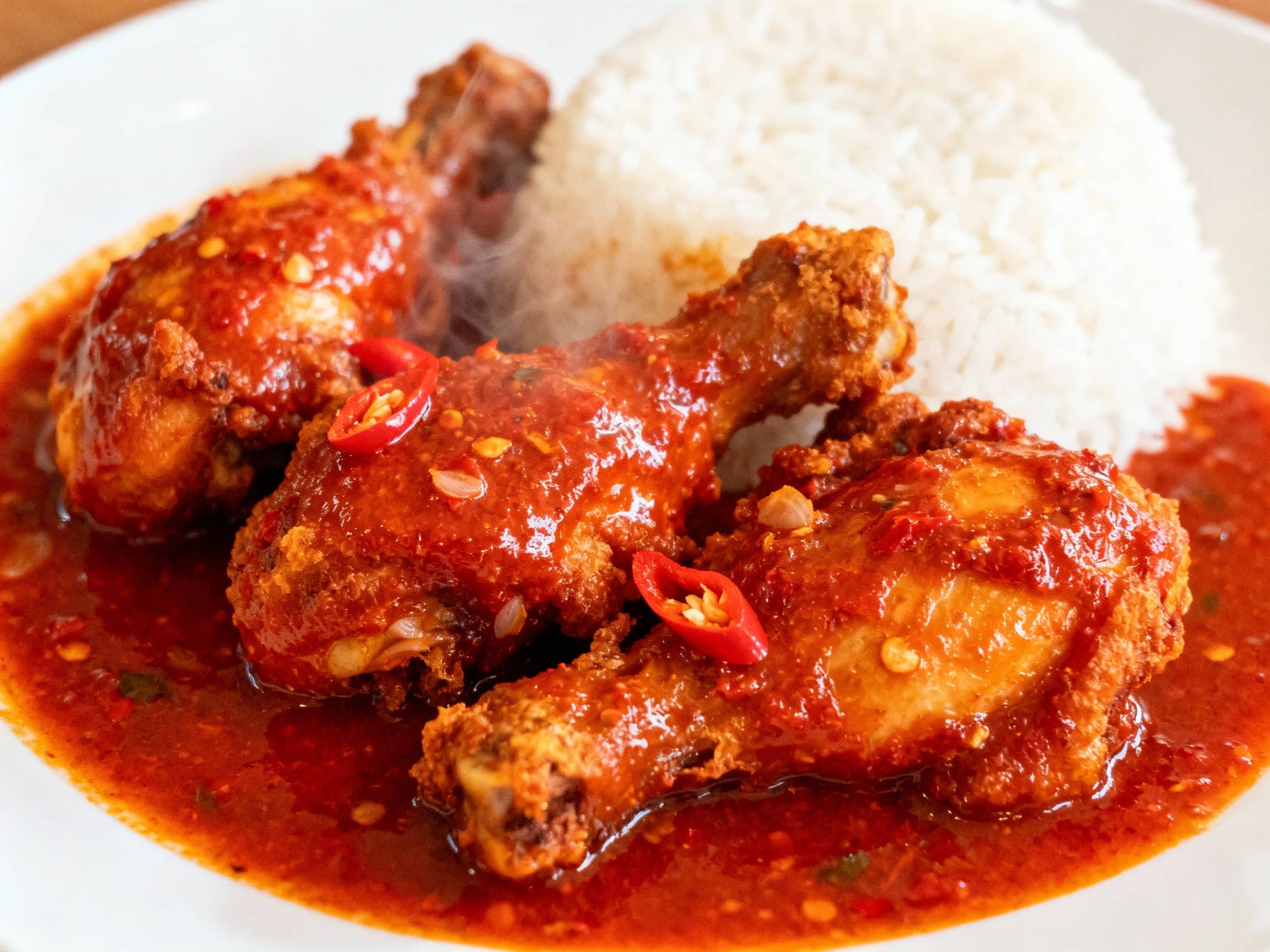 A mouth-watering close-up shot of 'Ayam Balado' (chicken in Balado sauce), viewed from a slightly high angle. The pieces of fried chicken are coated in a thick, glossy, vibrant red Balado sauce, glistening under warm overhead lighting. Small pieces of red chili and shallots are visible within the sauce. The dish is served on a simple white plate, with a serving of plain white rice to its side, emphasizing the sauce's intensity. The atmosphere is that of a bustling local Indonesian restaurant, with a hint of steam rising from the food.