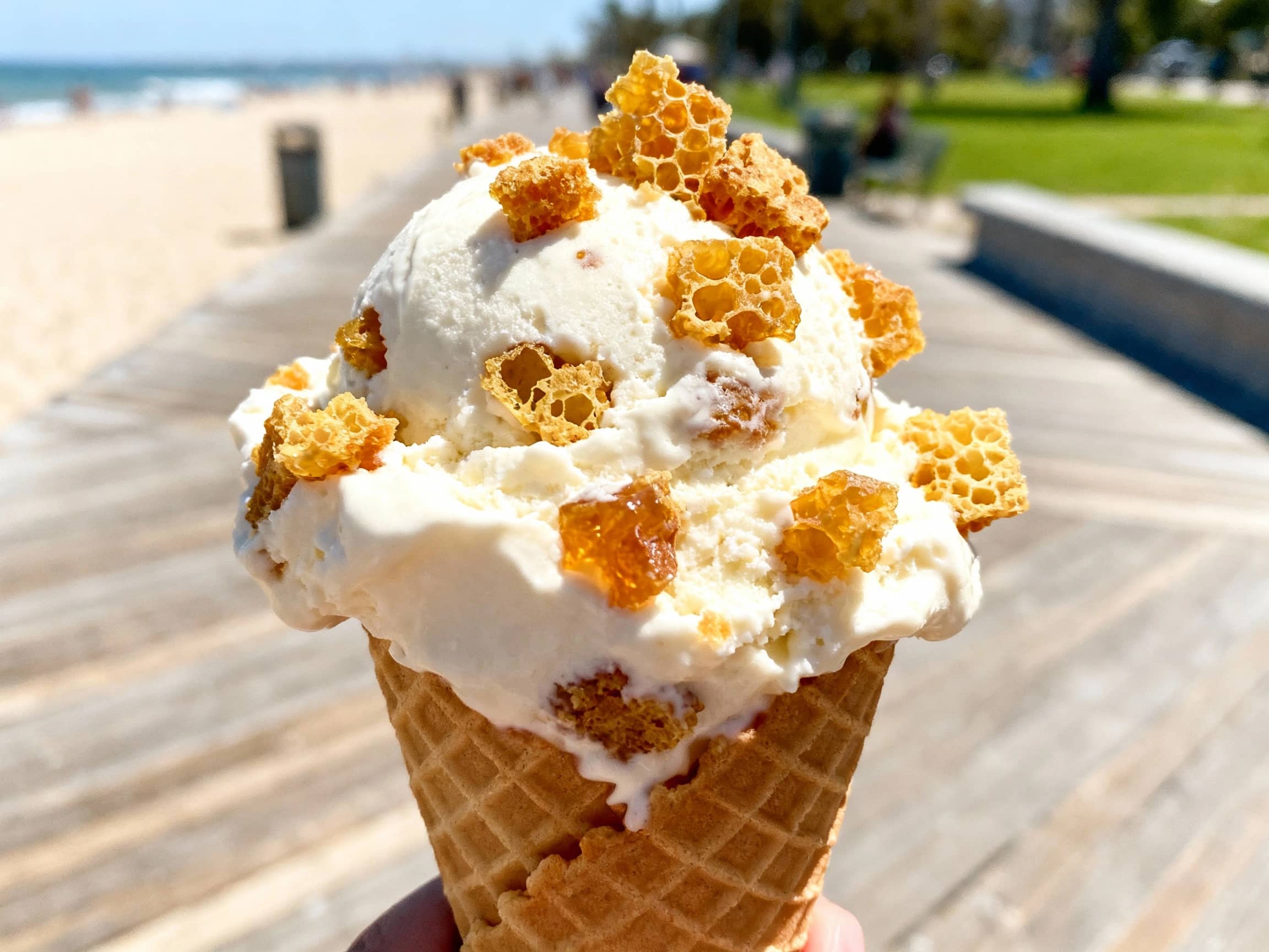 A close-up shot of Hokey Pokey ice cream being scooped into a waffle cone. The vanilla ice cream is speckled generously with golden-brown, irregularly shaped chunks of crunchy honeycomb toffee. The background shows a casual, outdoor setting, perhaps a beach boardwalk or a park on a sunny day. The lighting is bright and natural, highlighting the texture of the ice cream and toffee. The view is a tight close-up, emphasizing the delicious details. The atmosphere is one of simple, sweet, summery pleasure.