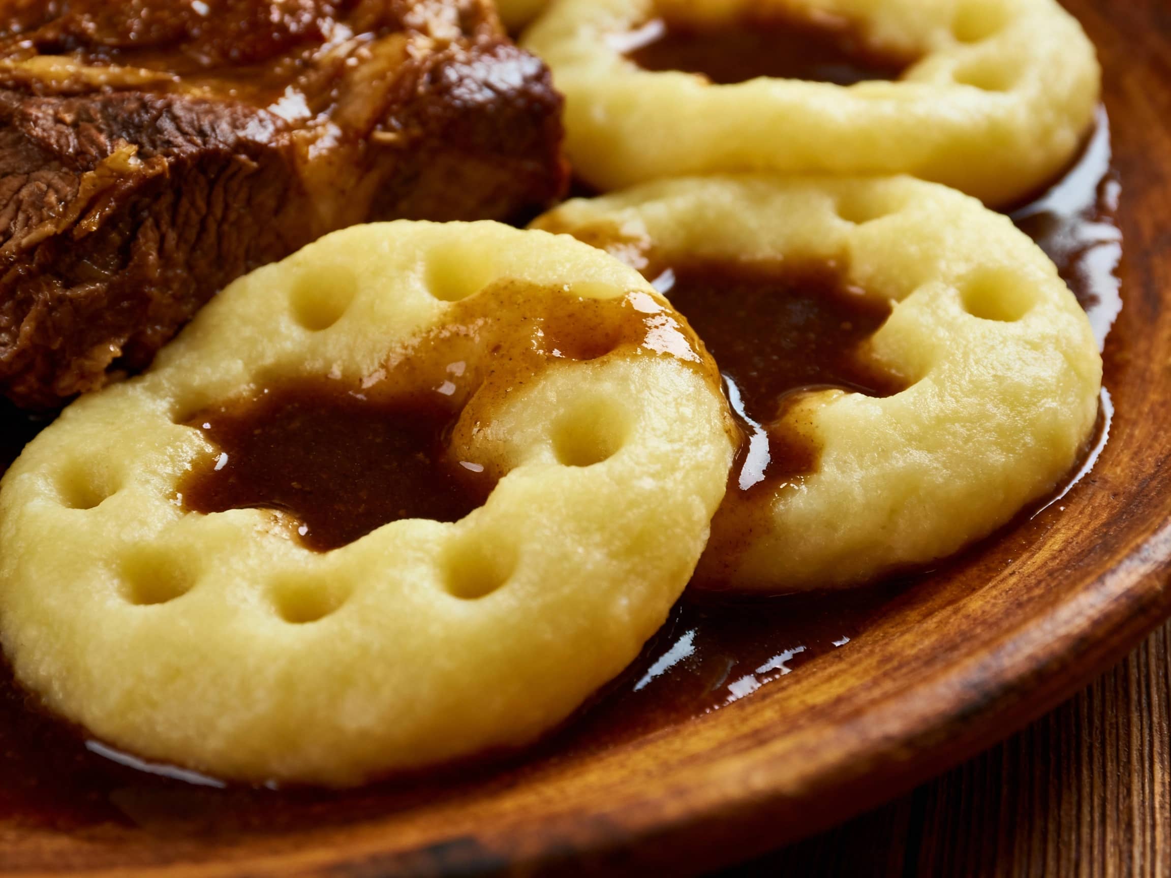 A close-up shot from a slightly elevated angle of several Kluski Śląskie, their characteristic dimple prominent, resting on a rustic wooden plate. Rich, dark gravy is pooled in the dimples and flowing around the dumplings. A succulent piece of braised meat sits nearby. The lighting is warm and inviting, highlighting the textures and inviting diners to enjoy this hearty Silesian classic.