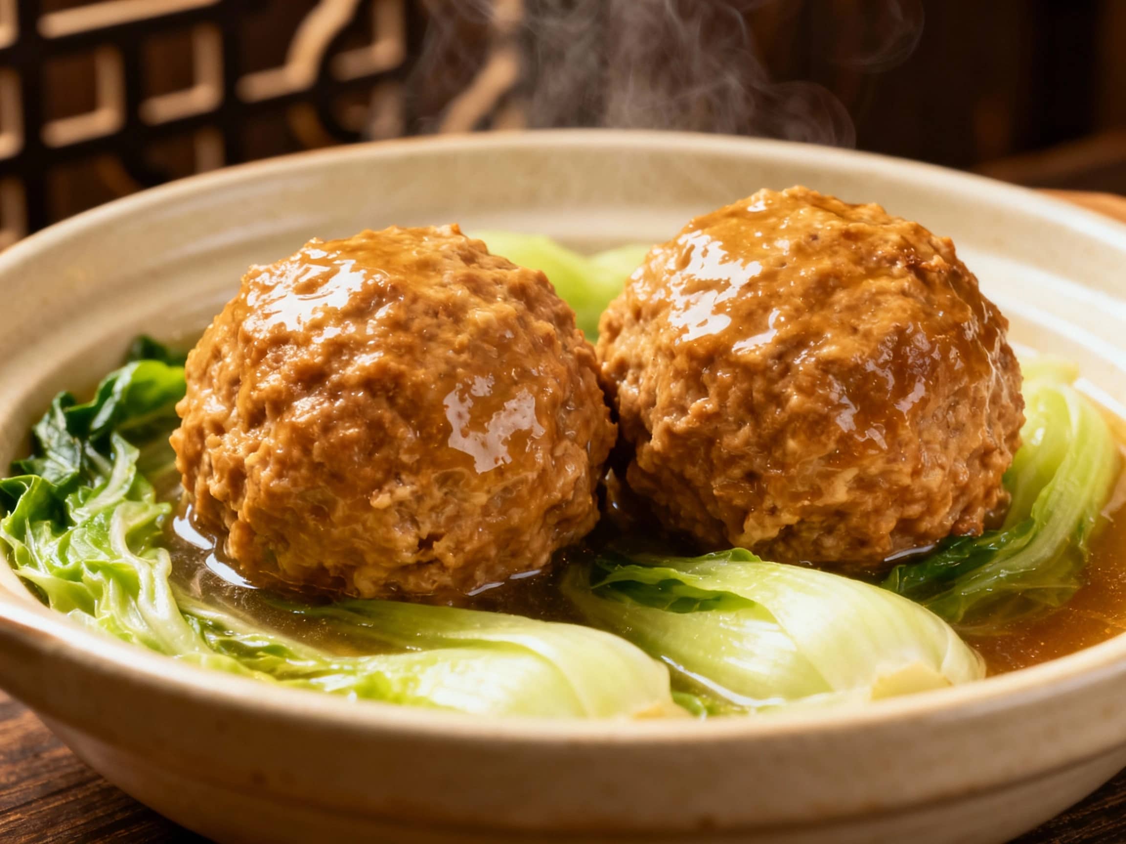 A close-up, eye-level shot of two large, tender Lion's Head Meatballs nestled in a shallow bowl atop a bed of wilted napa cabbage. The meatballs are a light, appealing brown, glistening with savory broth. The cabbage is a soft green, contrasting with the meatballs. Steam gently rises from the dish, indicating it's freshly served. The background is kept minimal and warm, possibly dark wood or a soft-focus traditional Chinese pattern, ensuring the focus remains on the comforting and hearty appearance of the meatballs. The overall impression is one of rustic elegance and a deeply satisfying meal.