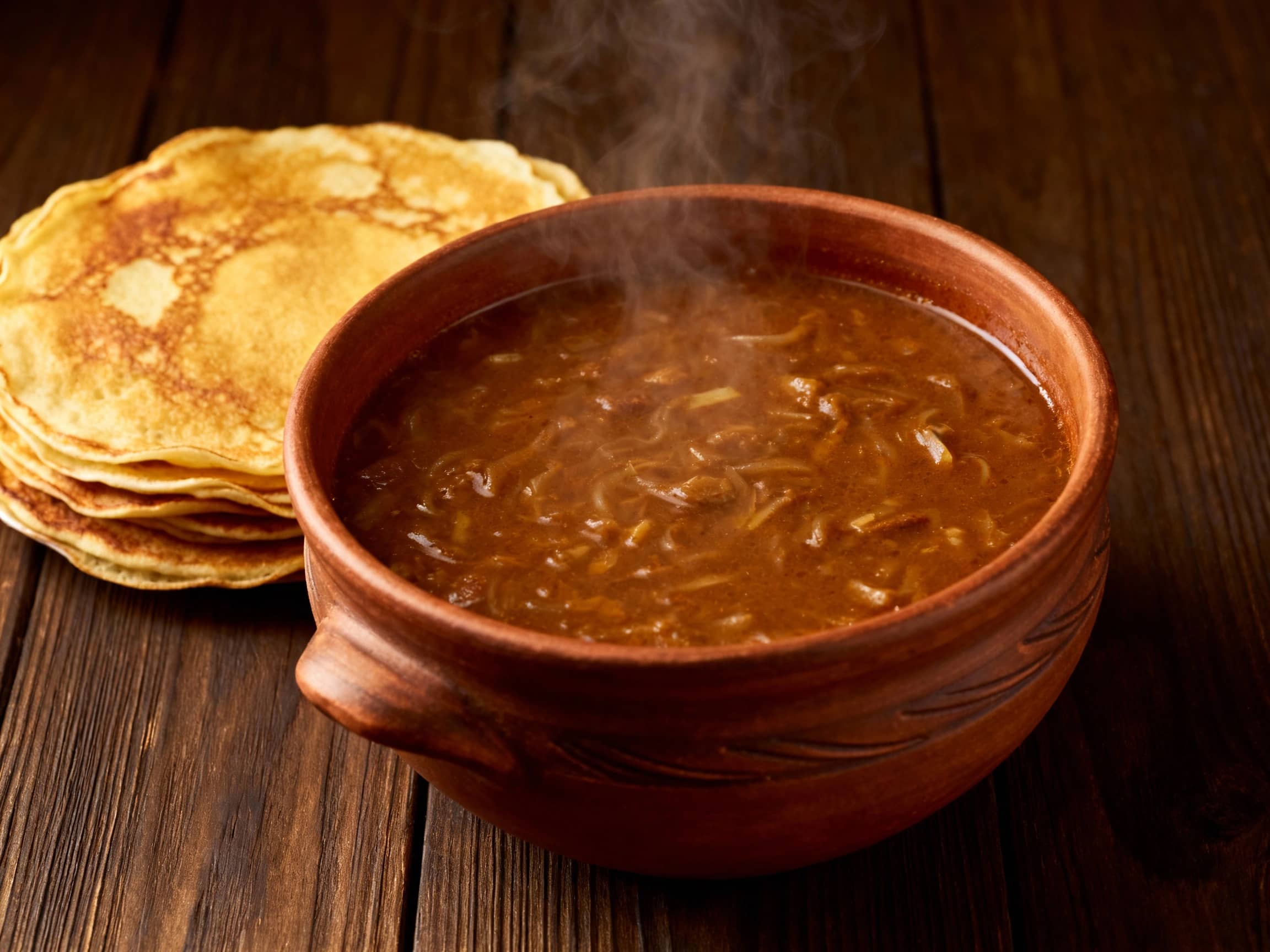 A rustic clay bowl filled with rich, brown Machanka, thick and steaming, served alongside a stack of golden brown, thin pancakes, on a dark wooden table, warm, inviting atmosphere, focused on the food, slightly overhead shot.