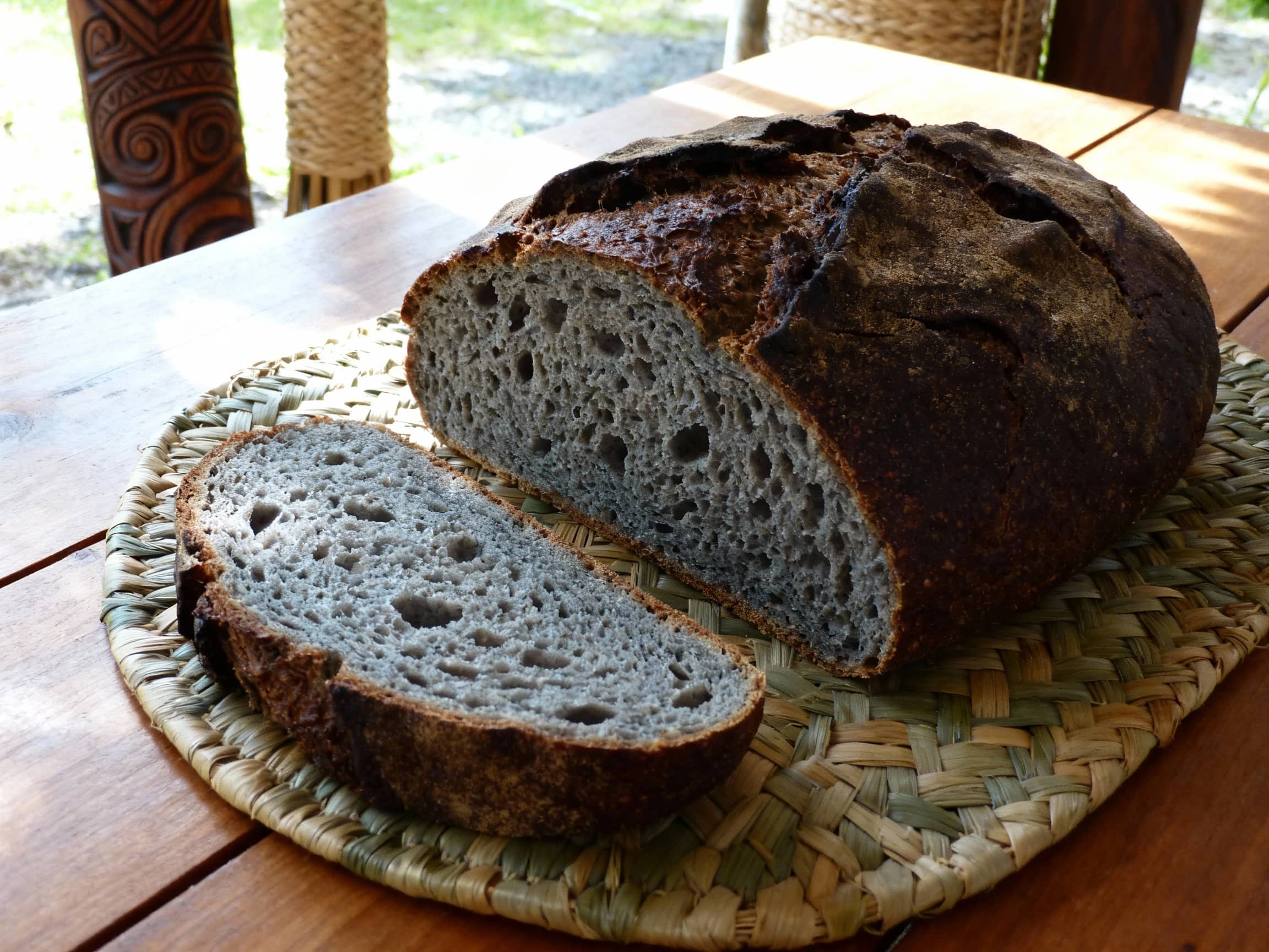 A rustic, dark-crusted loaf of Rewena Parāoa bread resting on a woven flax mat. The bread has a slightly irregular, dense shape, characteristic of sourdough. A slice has been cut, revealing a moist, open crumb with a slight greyish hue. It is placed on a simple wooden table, possibly in an outdoor setting with natural light filtering through. In the background, elements suggestive of Māori culture might be subtly present, like carved wood or woven materials. The view is a close-up, focused on the texture of the bread. The atmosphere is authentic and traditional.