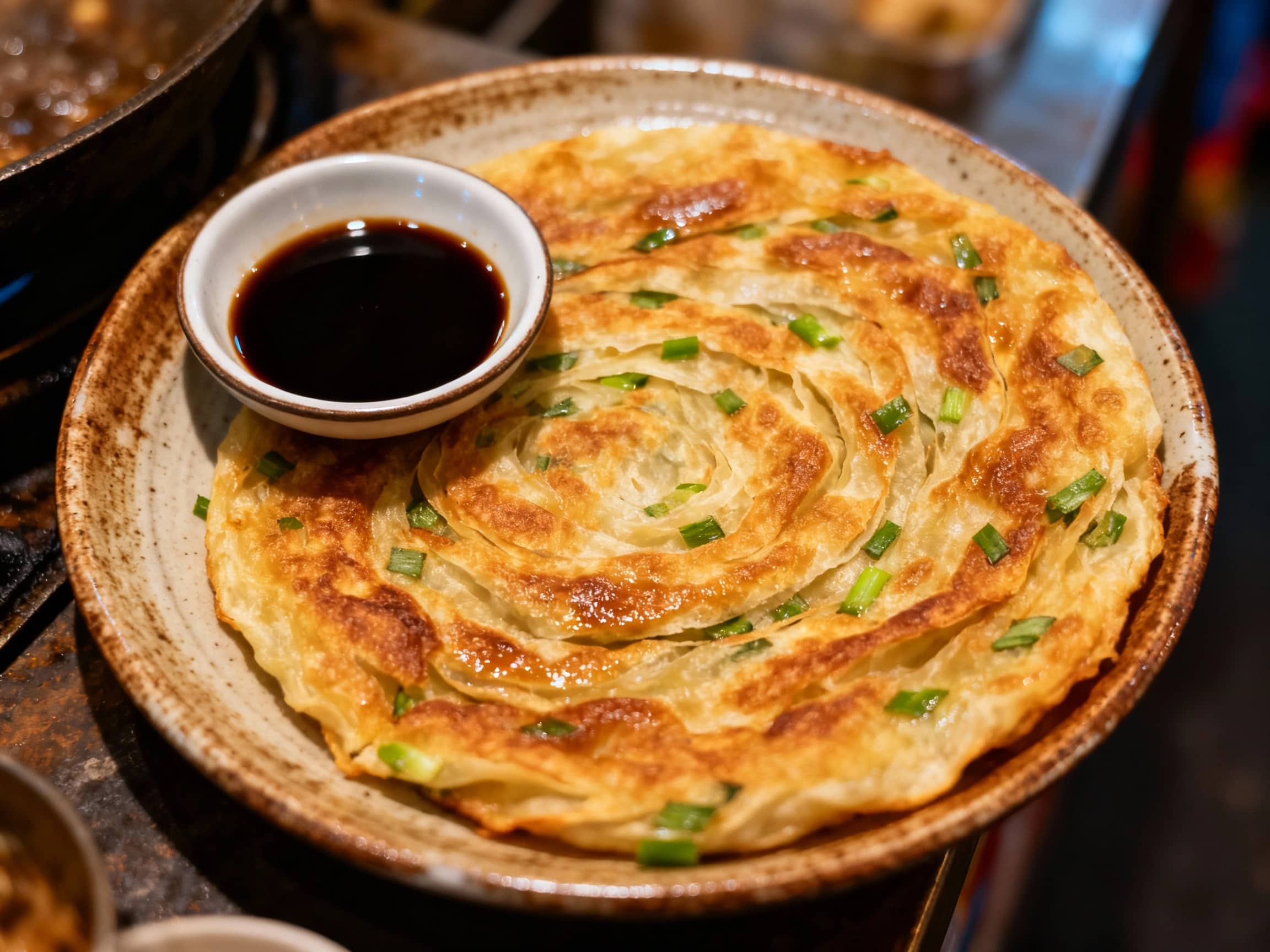 A close-up, top-down view of a freshly pan-fried, golden-brown scallion pancake on a rustic ceramic plate. The pancake is slightly oily, showing distinct layers and flecks of green scallions. A small dish of dark soy sauce-based dipping sauce sits to the side. The lighting is warm and inviting, suggesting a home-cooked meal or a bustling night market stall atmosphere. The background is slightly blurred, focusing attention on the texture and appeal of the pancake.