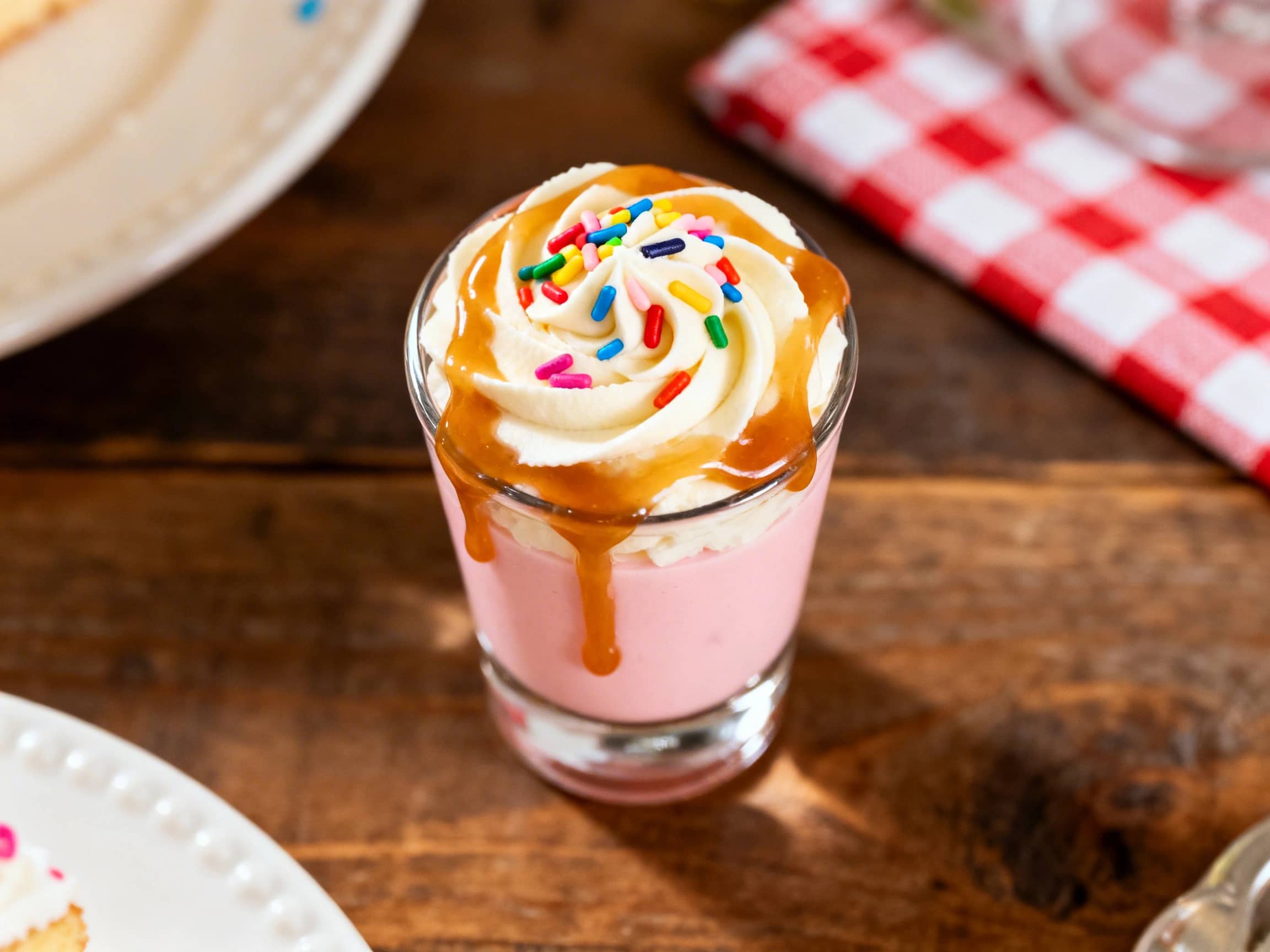 A single Birthday Cake Vodka Shot in a clear, small shot glass, presented against a soft, festive background. The creamy, pale yellow liquid fills the glass, and the rim is generously coated with bright, multicolored sprinkles. The overall presentation is inviting and celebratory, capturing the essence of a fun, dessert-themed beverage.