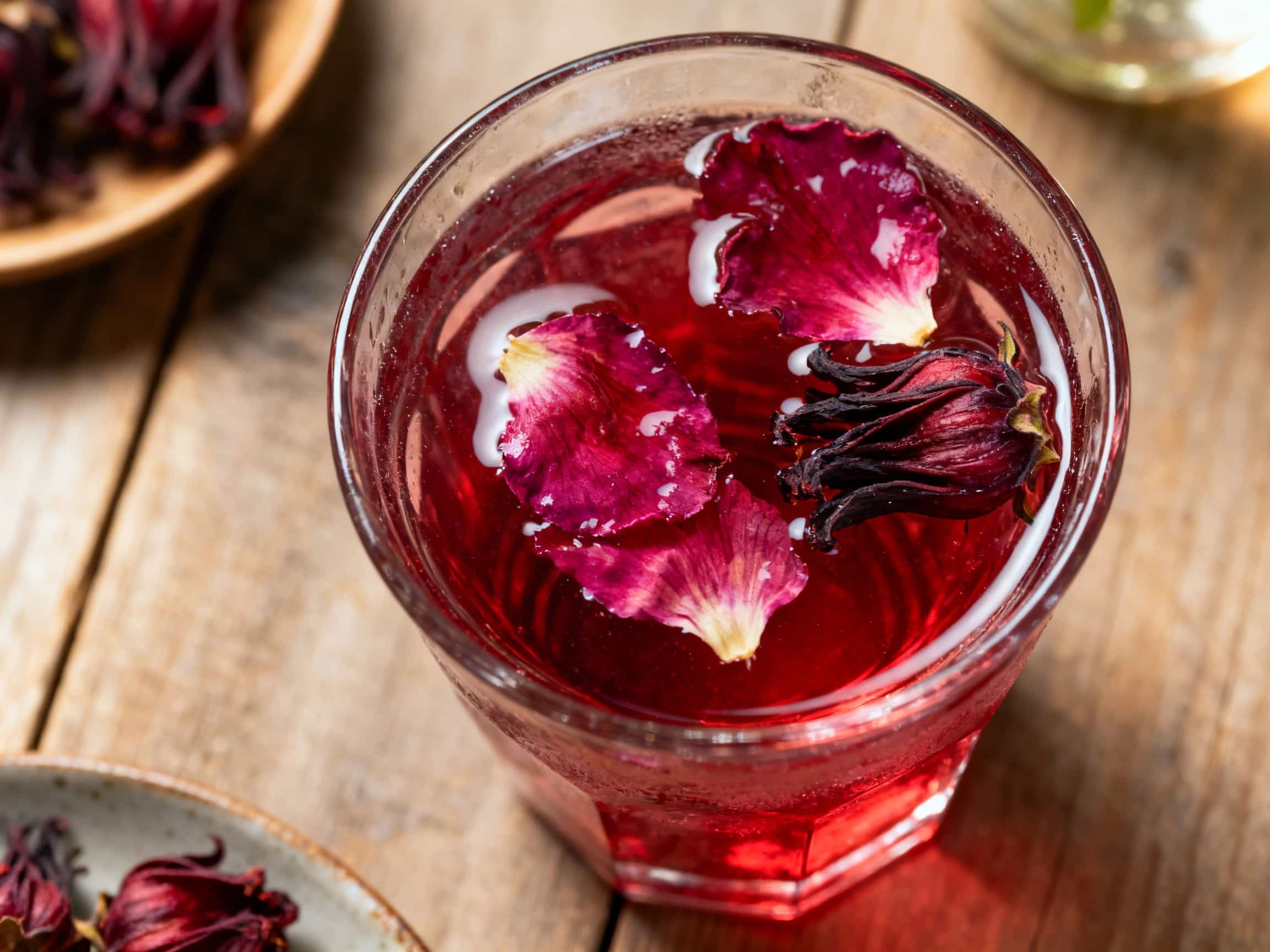 A beautiful deep crimson liquid, hibiscus and rose infused vodka, sits in a clear glass bottle on a rustic wooden table. Dried hibiscus flowers and rose petals are scattered around and within the bottle, creating an enchanting, artisanal display. The natural light highlights the rich color of the infusion, suggesting a vibrant and aromatic experience. It evokes the feeling of a sophisticated homem