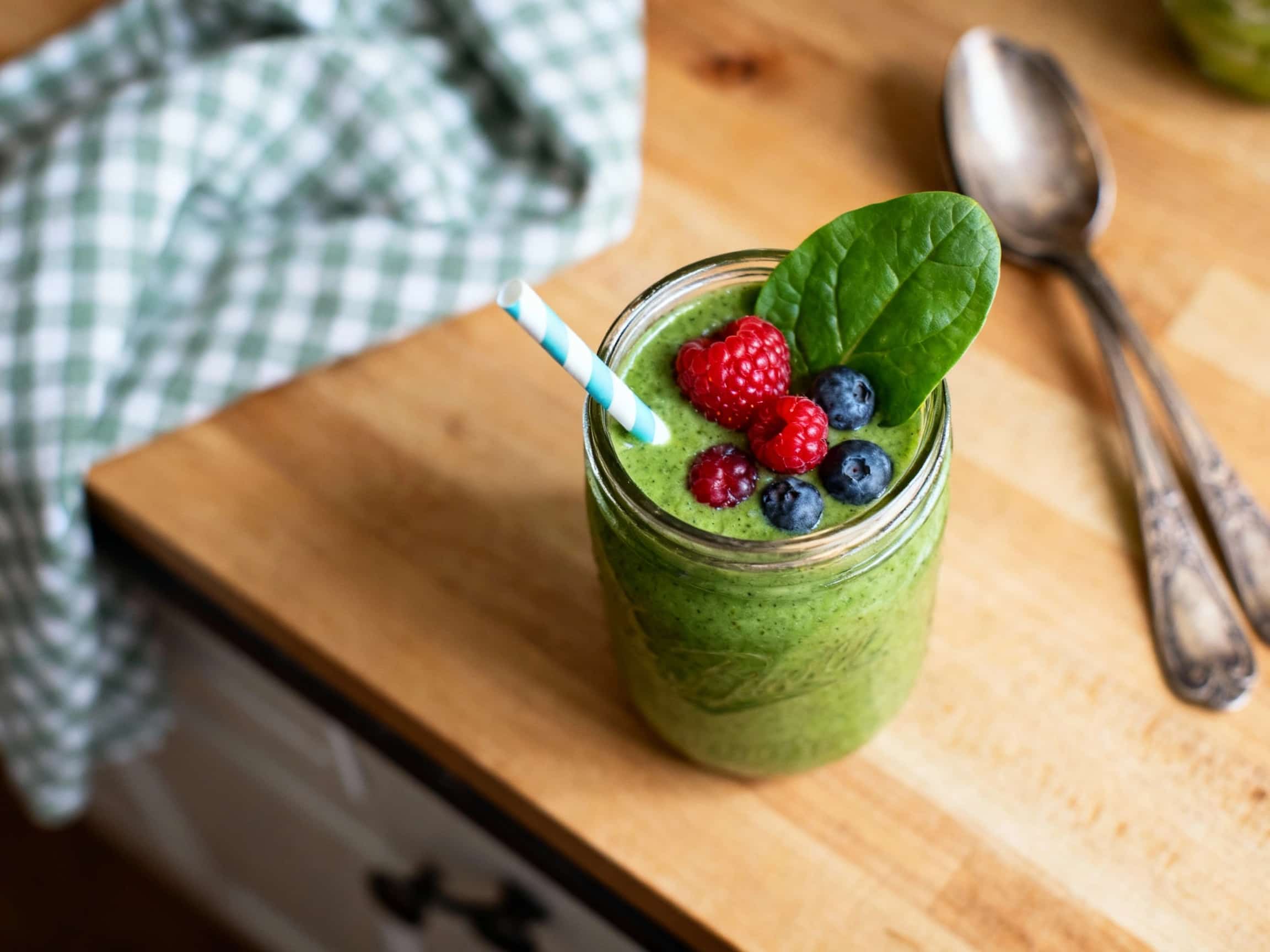 A vibrant magenta Berry Spinach Power Smoothie in a tall, clear glass. The smoothie is topped with fresh raspberries and a sprinkle of chia seeds, creating an appealing contrast. The glass is placed on a light wooden surface, with a soft, sunlit background that suggests a healthy, energizing start to the day. The drink's rich color and garnishes highlight its fresh, nutritious ingredients and invi