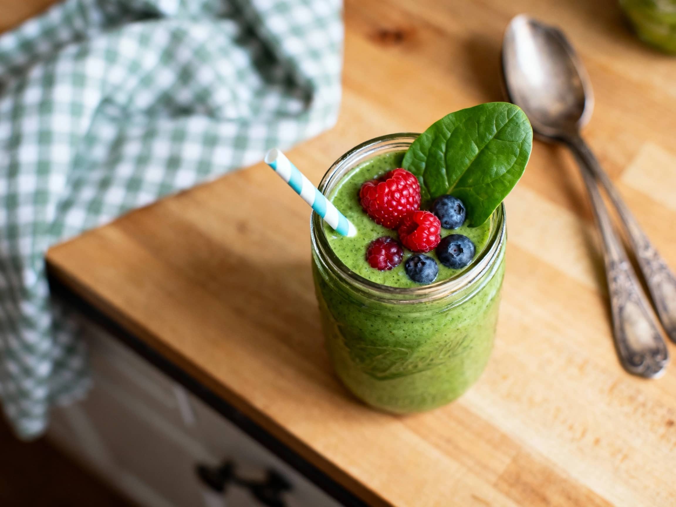 A vibrant magenta Berry Spinach Power Smoothie in a tall, clear glass. The smoothie is topped with fresh raspberries and a sprinkle of chia seeds, creating an appealing contrast. The glass is placed on a light wooden surface, with a soft, sunlit background that suggests a healthy, energizing start to the day. The drink's rich color and garnishes highlight its fresh, nutritious ingredients and invi