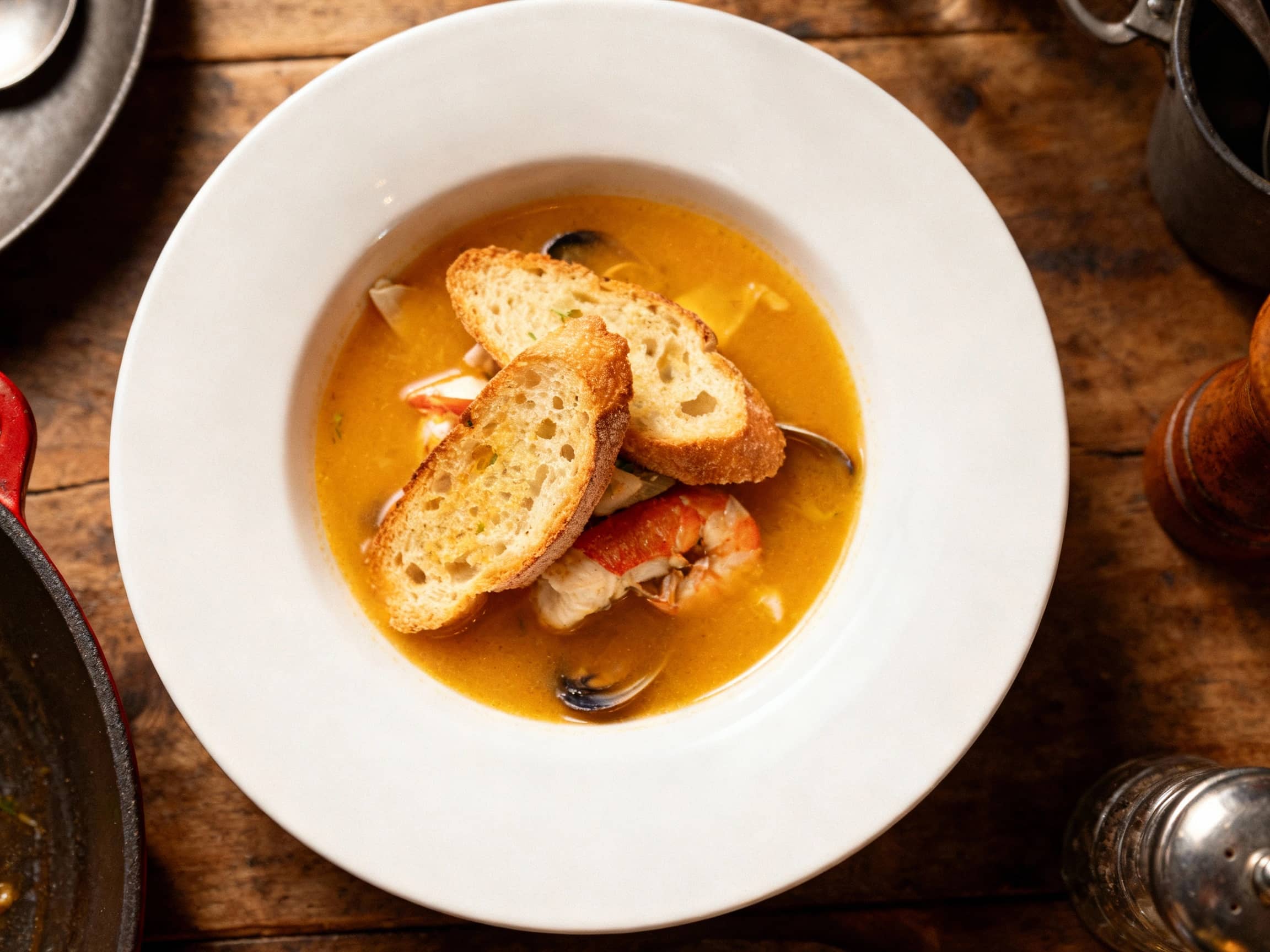 Miniature servings of rich, saffron-infused bouillabaisse, brimming with colorful seafood, are presented in small glasses alongside golden toasted crostini, offering a sophisticated appetizer.