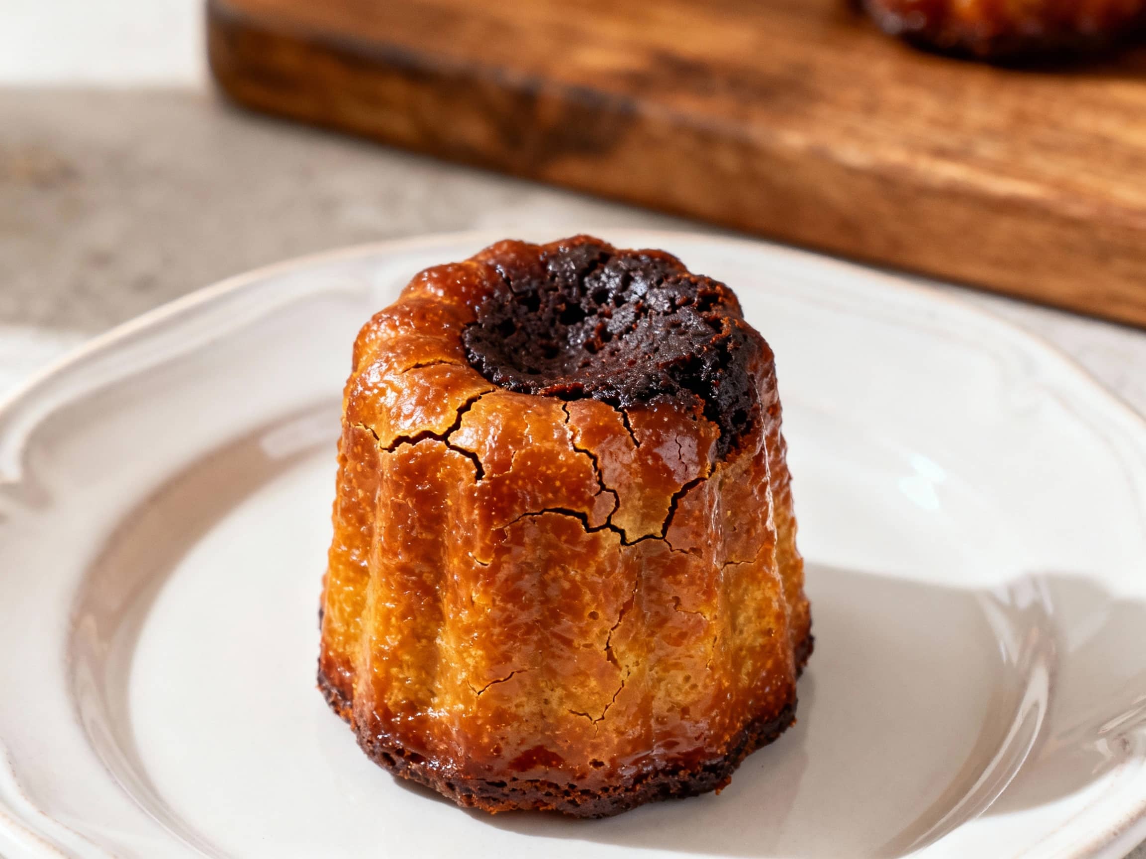 Golden-brown Bordeaux cannelés with a tall, caramelized crust and a custardy, dark interior sit attractively arranged on a rustic wooden board, ready to be enjoyed.