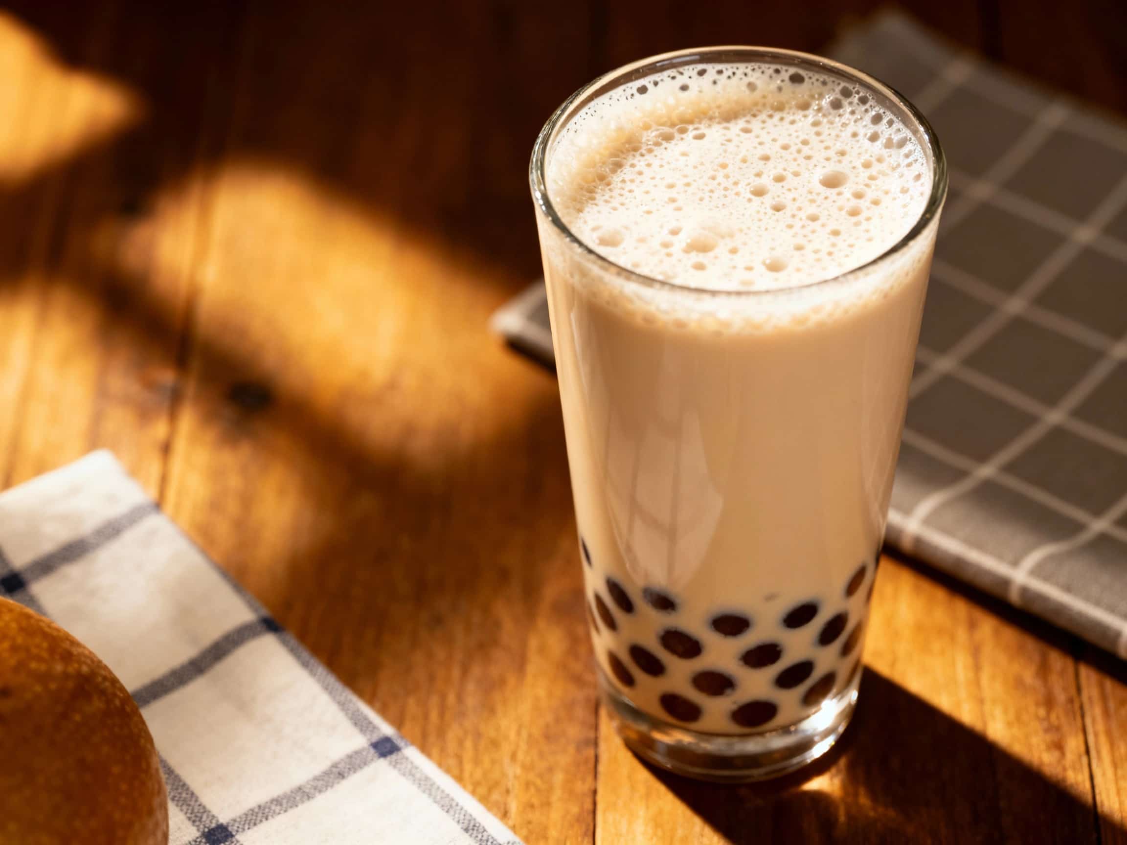 A tall glass of creamy brown pearl milk tea, topped with a frothy layer of foam and a generous scattering of dark tapioca pearls, is ready to be enjoyed.