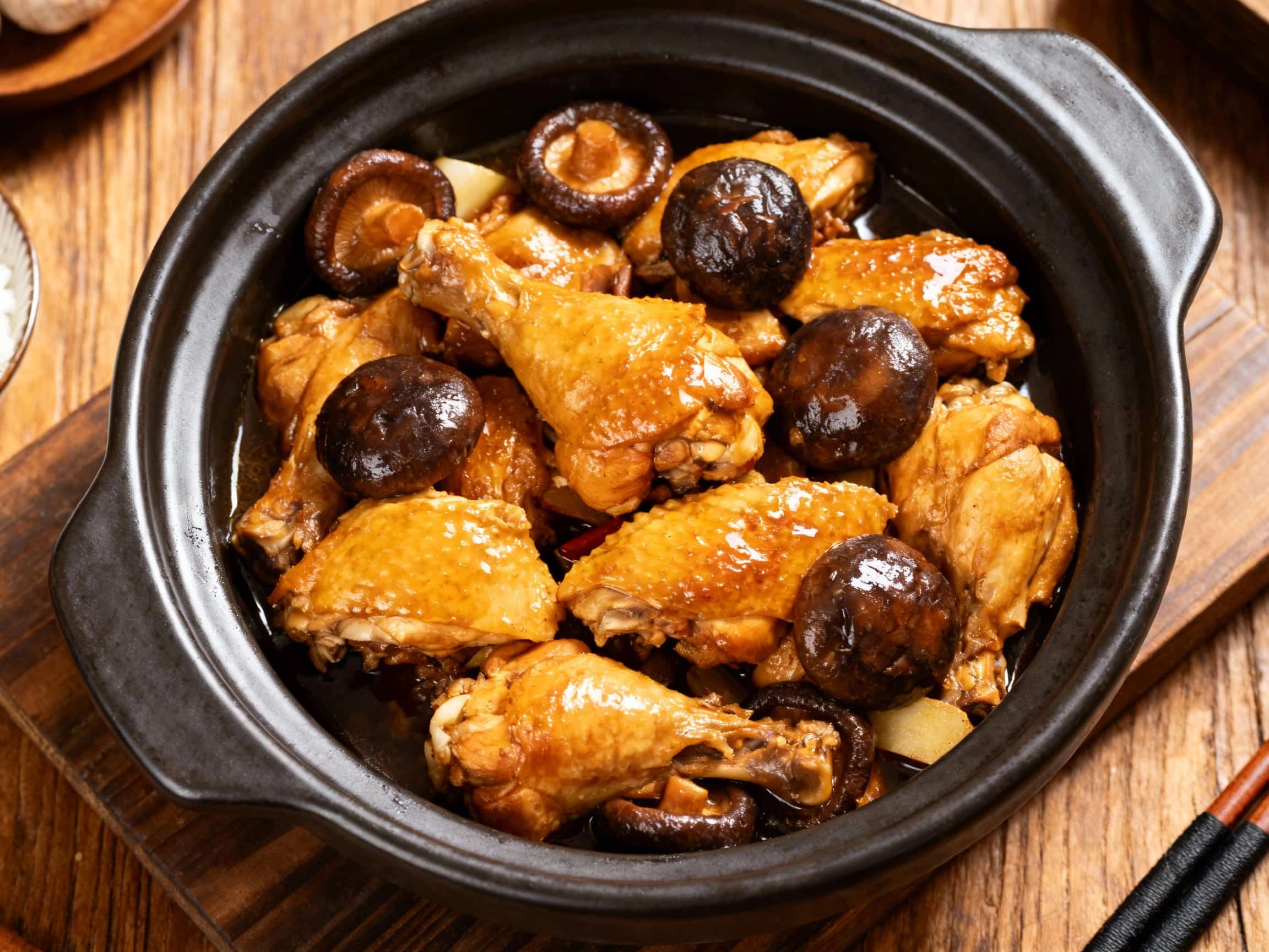 Tender, dark chicken pieces glazed in a rich, glossy three-cup sauce sit atop fluffy white rice in a rustic clay pot, garnished with fresh cilantro and sliced shiitake mushrooms.