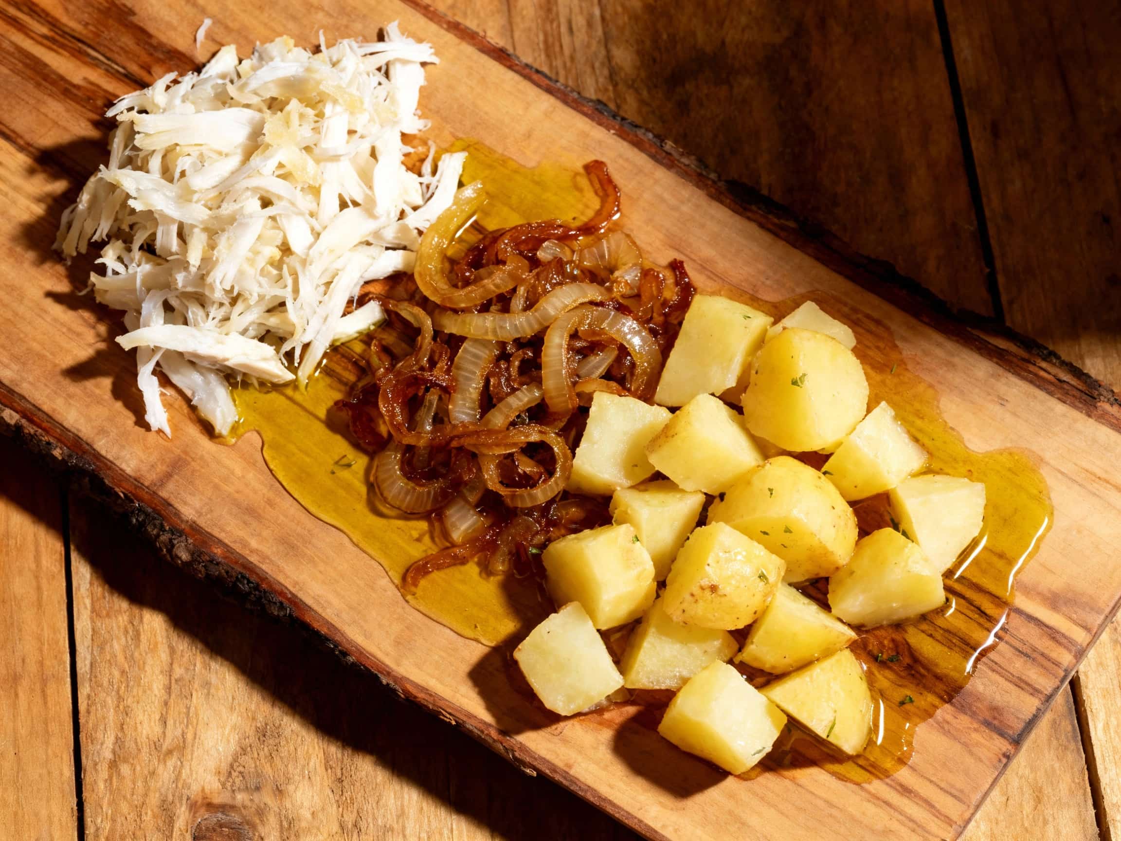 Flaky white cod, potatoes, and onions are artfully arranged on a white plate, drizzled with olive oil and garnished with black olives and a sprinkle of parsley.