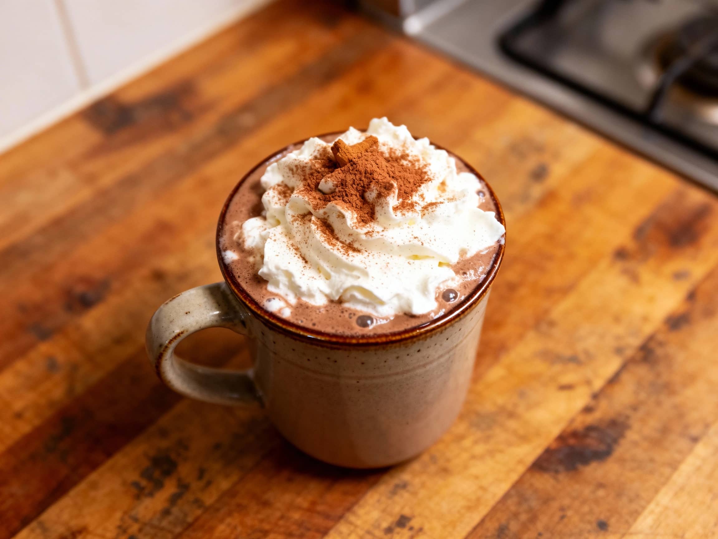 A close-up of a dark, rich Mexican mocha hot chocolate in a ceramic mug, with visible steam rising. It's topped with a swirl of white whipped cream and sprinkled with cinnamon. The mug rests on a dark wooden surface in a cozy, softly lit environment, conveying warmth and indulgence.