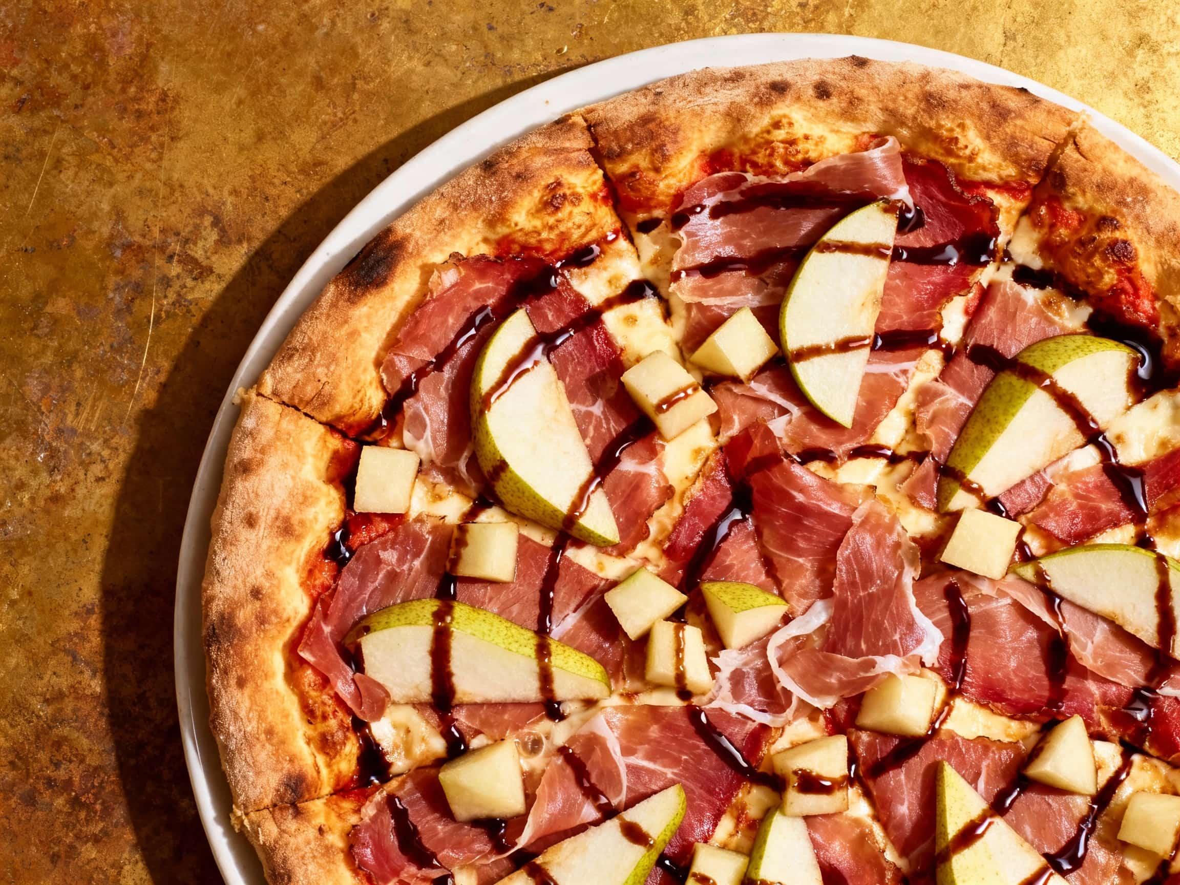 A close-up of a square slice of Prosciutto and Pear Sicilian Pizza on a wooden board. The pizza features a thick, focaccia-like crust, topped with melted mozzarella, thinly sliced prosciutto, and sweet caramelized pear. A dark balsamic glaze is drizzled over the pizza, and fresh green arugula leaves are scattered on top, creating a vibrant and appetizing appearance.