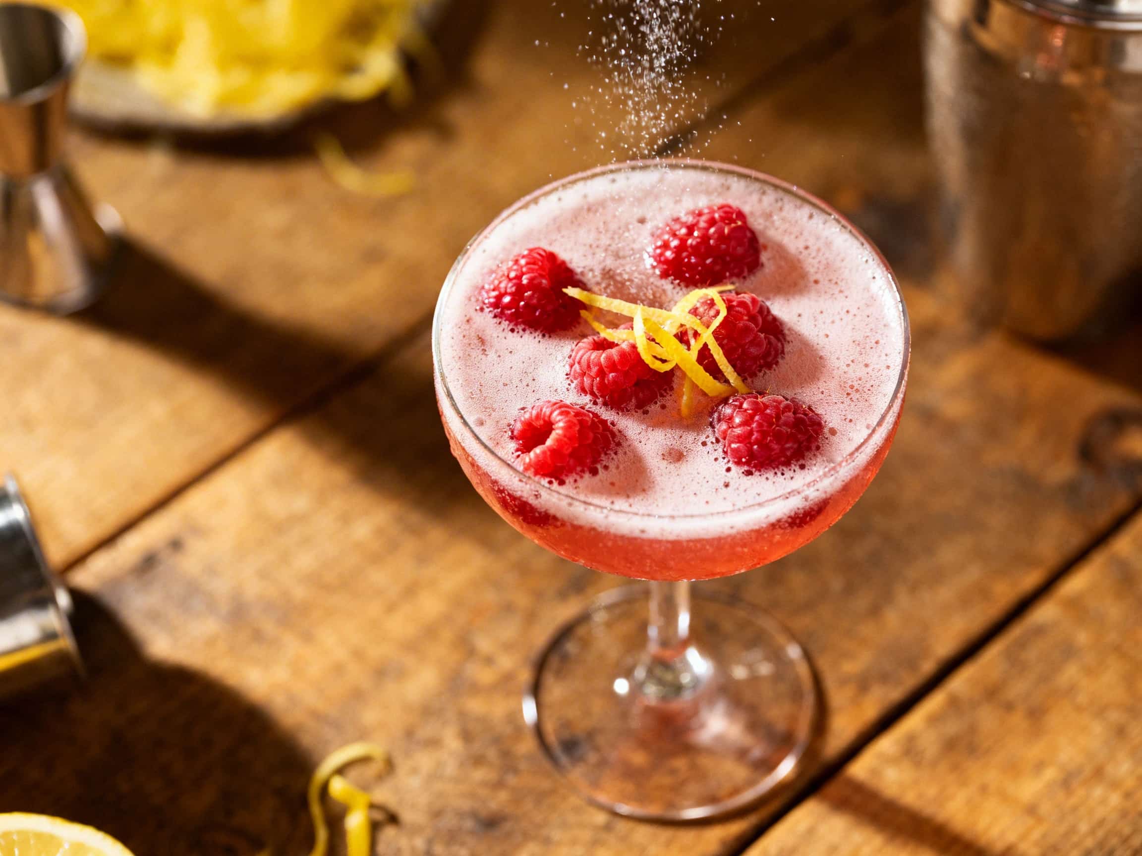 A beautiful deep ruby-red raspberry limoncello sparkler shot fills a small, clear shot glass, garnished with a fresh green mint sprig and two ripe red raspberries. The drink appears effervescent with visible bubbles, suggesting a light, refreshing taste. It's presented on a dark, elegant surface under warm, inviting light, setting a festive and sophisticated mood for a celebratory drink.