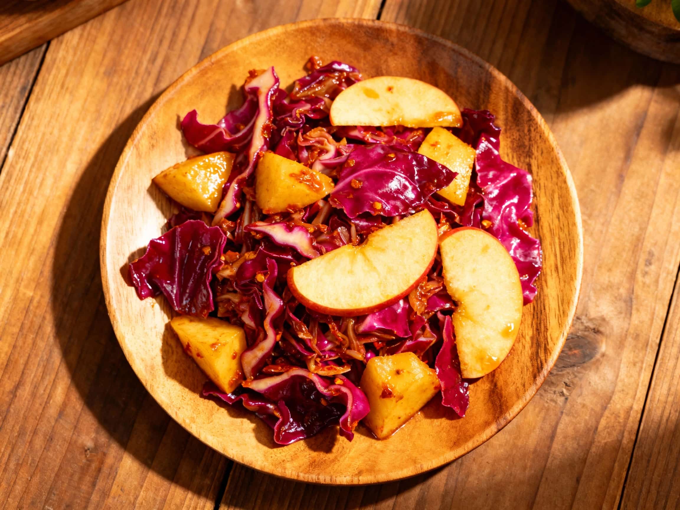 An overhead shot of a rustic wooden bowl brimming with bright red and white spicy fermented cabbage and apple. The slaw is finely shredded, showing off visible crimson chili flakes, delicate slices of red apple, and thin slivers of fresh ginger, all glistening in a light brine. The bowl sits on a natural wooden surface under soft, diffuse lighting, creating an inviting and wholesome visual.