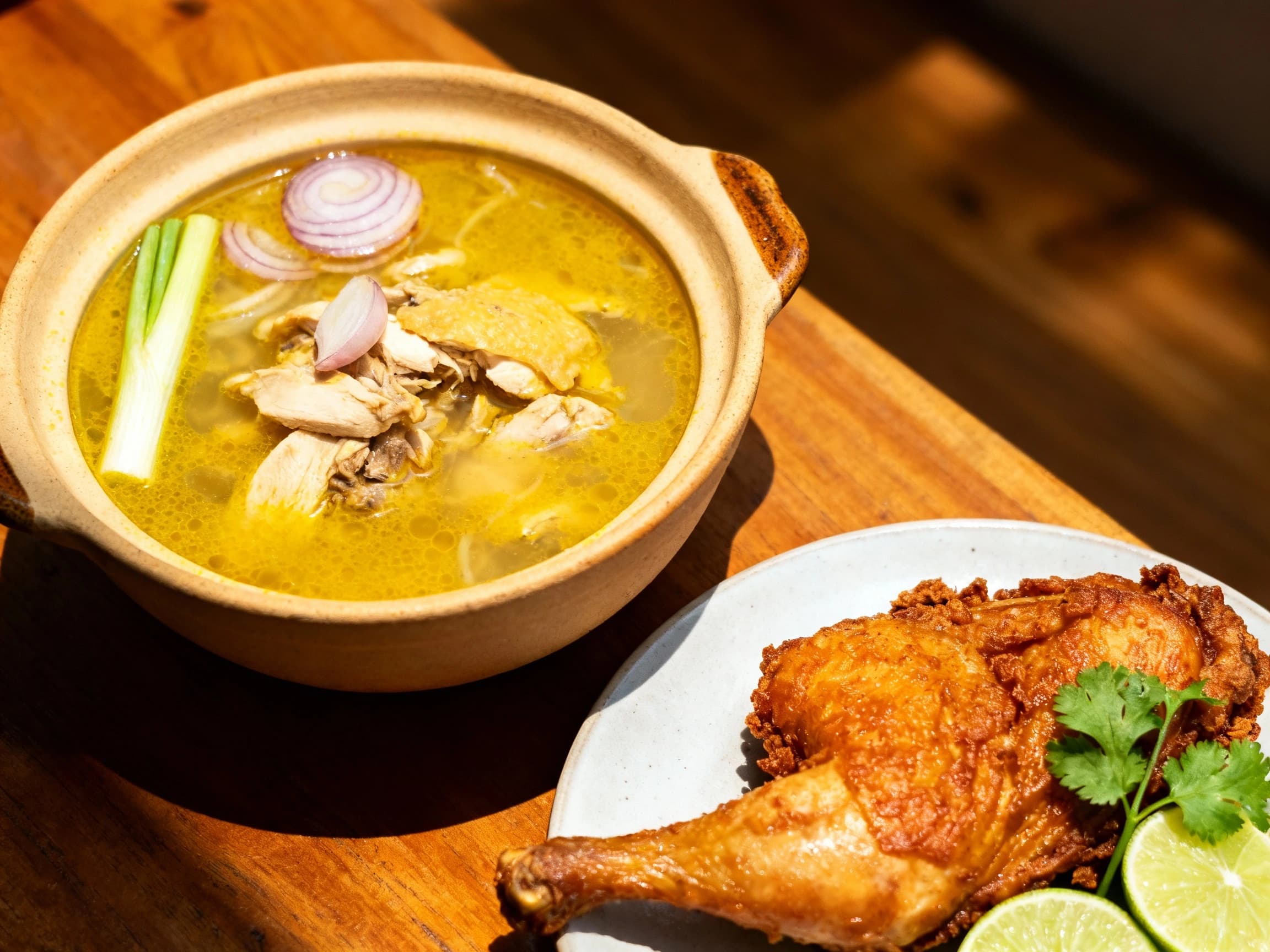 A steaming bowl of Indonesian Soto Ayam, featuring golden crispy fried chicken pieces arranged alongside delicate rice vermicelli noodles and fresh garnishes. The savory turmeric-infused broth is poured over the ingredients, creating an inviting and aromatic presentation perfect for a traditional meal.